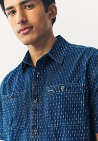 Chemise bleu marine à boutons avec un motif texturé de lignes blanches verticales, deux poches avant et des boutons marron. Manches courtes, col classique.