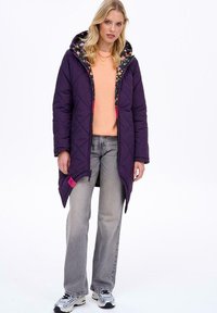 Prošívaný fialový vnější kabát s vzorovanou černou kapucí, růžovými akcenty a předním zipem. Nositelný přes světle oranžový top a šedé džíny.