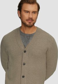 Beige Strickjacke mit V-Ausschnitt, strukturiertem Stoff, fünf schwarzen Knöpfen an der Vorderseite und rippenartigen Akzenten entlang des Kragens und der Bündchen.
