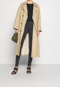 Abrigo largo beige con grandes solapas y botones marrones, combinado con jeans ajustados negros y sandalias de tacón negras. Sosteniendo un bolso verde.