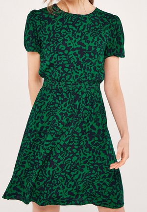 Femme portant une robe verte à manches courtes avec des motifs abstraits noirs et de feuilles, une taille élastique et une jupe légèrement évasée.