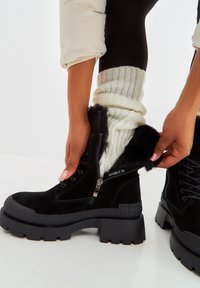 Botas de tobillo de ante negras con una suela gruesa y texturizada, detalles de piel de animal alrededor del tobillo y un zipper lateral. Combinadas con un calcetín de punto crema.
