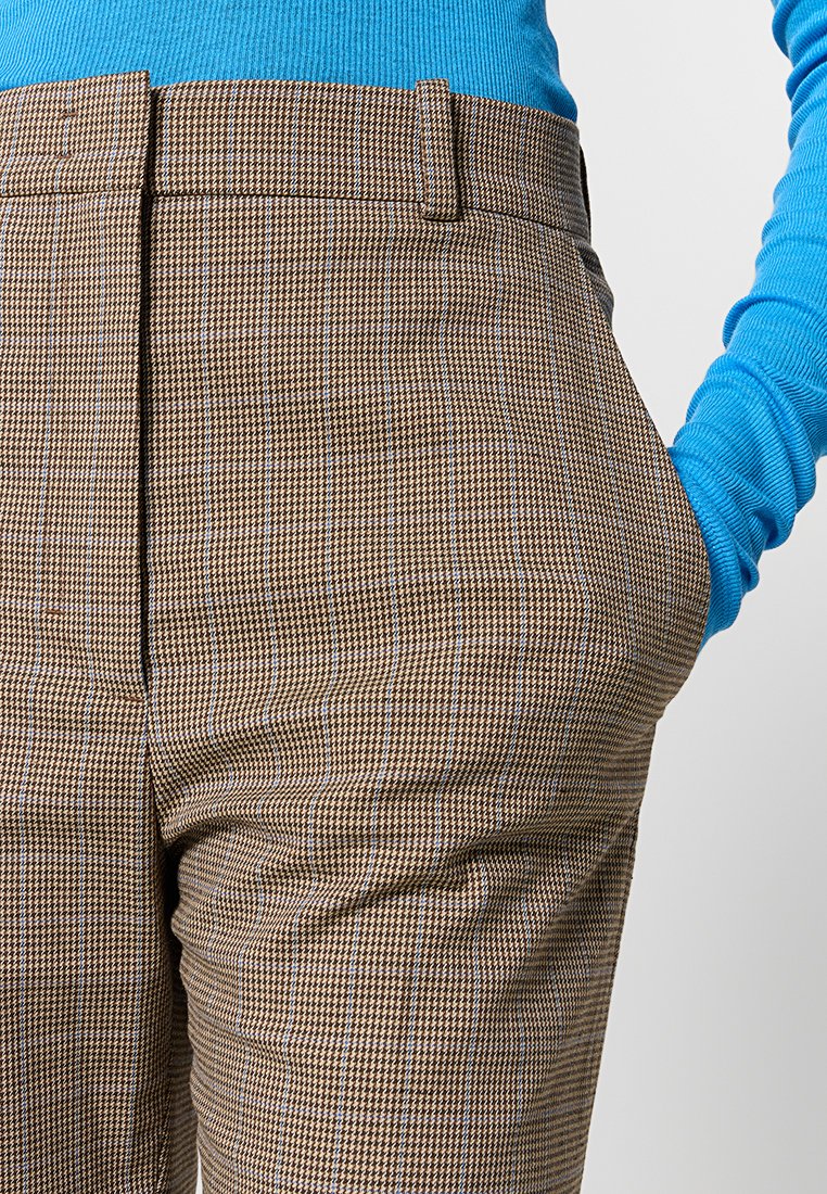 Brązowe spodnie w kratę z drobnym wzorem, z płaskim przodem i bocznymi kieszeniami, noszone z niebieską, prążkowaną bluzką z długim rękawem.