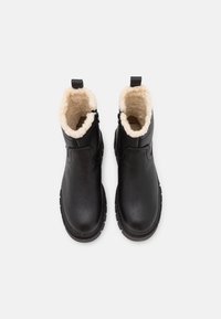 Svarta ankelboots med en kornig läderexteriör, fluffigt vitt foder och texturerad gummisula. Ovanifrån syns de rundade kanterna och dragfliken.