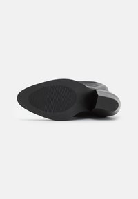 Černá podrážka boty na vysokém podpatku s hladkým zaobleným tvarem, strukturovaným gumovým povrchem a vodorovnými drážkami pro trakci. Zvednutý podpatek pro oporu.