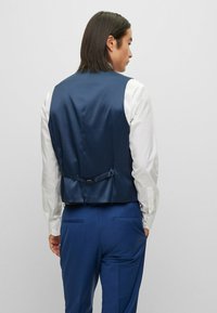 Marineblaues Weste aus glattem Stoff, strukturierter Schnitt, verstellbarer Rückenriemen, kombiniert mit einem weißen Hemd und blauen Hosen.