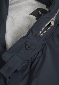 Dark navy jacket with silver zipper partially unzipped, buttoned strap detail, and light gray inner lining with visible black clothing label.