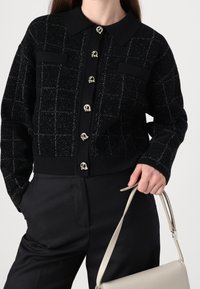 Giacca cropped a quadri neri con accenti di bottoni argentati, dotata di colletto e due tasche frontali. Il materiale ha una finitura leggermente testurizzata.