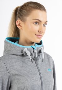 Grå zip-jacka med huva, med en blå innerv lining och flätade vita dragband. Inkluderar en liten blå logotyp på bröstet.