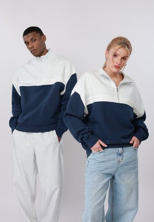 To modeller iført matchende sweatshirts med halvlange lynlåse i marineblå og lysegrå farveblokke; den ene iført lysegrå joggingbukser, den anden i lyseblå jeans.