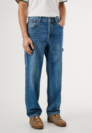 Homme portant un jean bleu ample avec la main dans la poche et des baskets marron, debout devant un fond uni.