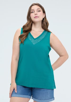 Vrouw met lang bruin haar draagt een turquoise mouwloos topje met strass detail en spijkershorts, staand tegen een lichte achtergrond.