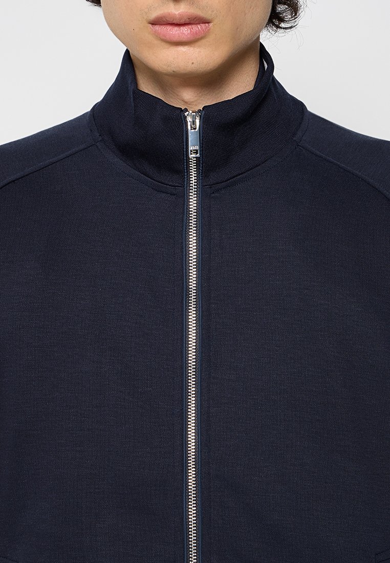 Granatowa kurtka zip-up z wysokim kołnierzem, miękka tkanina i srebrny zamek błyskawiczny. Charakteryzuje się eleganckim, minimalistycznym designem i rękawami raglanowymi.