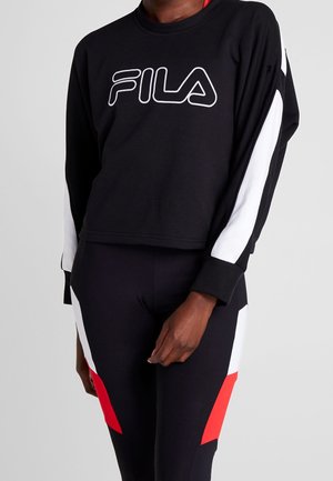 Persoon draagt een zwarte FILA sweater met witte mouwstrepen en bijpassende zwarte leggings met witte en rode geometrische panelen.