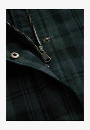 Kurtka w kratę w odcieniach ciemnej zieleni, z metalowym zamkiem błyskawicznym i ozdobnymi guzikami. Tkanina ma teksturowaną powierzchnię z nakładającymi się kwadratami.