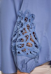 Blusa blu chiaro con dettagli in pizzo intricati sulle maniche e una finitura testurizzata. Le maniche sono arricciate ai polsi.