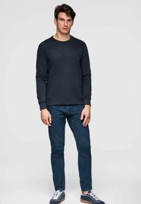 Felpa blu navy con logo "OMBRE" in design scuro, maniche lunghe e scollo a girocollo, abbinata a jeans in denim aderenti e sneakers blu navy.