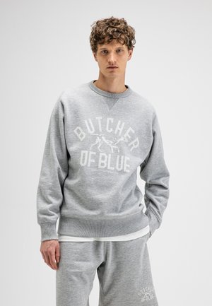 Junger Mann mit lockigem Haar, der einen hellgrauen Pullover und passende Jogginghose mit dem Schriftzug „Butcher of Blue“ und einem Hundemotiv trägt.