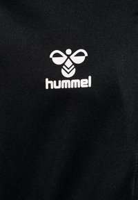 Musta kangas, jossa on valkoinen Hummel-logo. Logossa on tyylitelty mehiläisdesign, jossa on geometrisia muotoja ja pehmeitä viivoja.