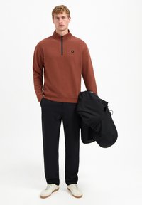 Man in roestkleurige kwart-rits trui, zwarte broek, witte sneakers, die een zwarte jas vasthoudt tegen een effen witte achtergrond.