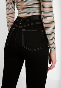 Svarta jeans med vit sömnad, hög midja och två bakfickor, i kombination med en randig långärmad topp. Märket syns vid midjebandet.