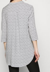 Kobieta w długiej, wzorzystej tunice w kolorze białym i czarnym z rękawami o długości trzech czwartych, stojąca tyłem do kamery.