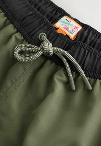 Maslinasto zelene kupaće gaće s crnim elastičnim pojasom, koje imaju pertlu s sivim naglascima i šareni znak s natpisom "NEXT Swimwear."