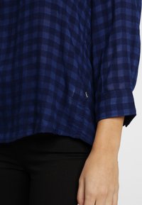 Blå och svart rutig långärmad skjorta på person som bär svarta byxor, med armen och handen synlig vid sidan.