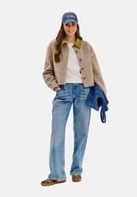 Beżowa krótka kurtka z zielonym kołnierzem, biała sweter, jasnoniebieskie dżinsy i mokasyny w panterkę. Trzyma niebieską torebkę z zamszu.