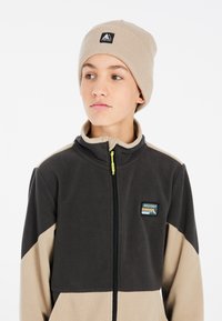 Fleece jack in zwart en beige met een hoge kraag, ritssluiting aan de voorkant en logopatch. Vergezeld van een tan geknoopte muts.