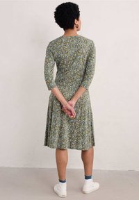 Sukienka w kwiaty w zielono-niebieskim kolorze z rękawami do łokci, dopasowanym biustem, rozkloszowanym dołem i białymi trampkami. Tło gładkie i jasne.