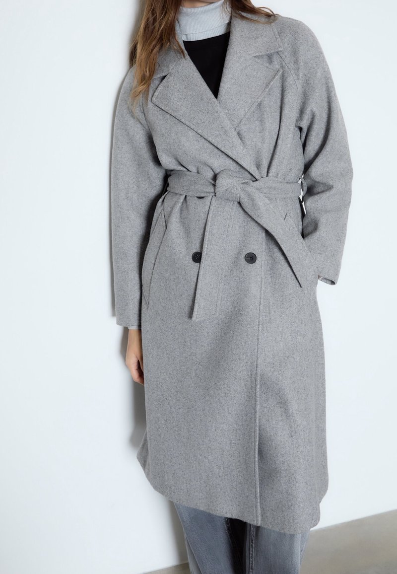 Femme portant un manteau en laine gris ceinturé, arrivant aux genoux, par-dessus un haut à col roulé noir et gris clair, debout contre un mur blanc.