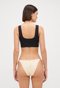 Juodas trumpas bikini viršus su plačiomis petnešėlėmis, derinamas su šviesiai beigėmis tekstūruotomis bikini kelnaitėmis su šoninėmis užrišimais ir dekoratyvinėmis karoliukais.