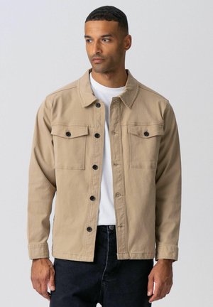 Mand iført beige knappet jakke over hvid skjorte og sorte bukser, stående mod en ensartet lysegrå baggrund.