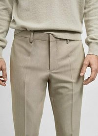 Pantalons beige ajustés avec un devant plat, une texture subtile, des passants de ceinture et une coupe droite. Portés avec un pull de couleur claire.