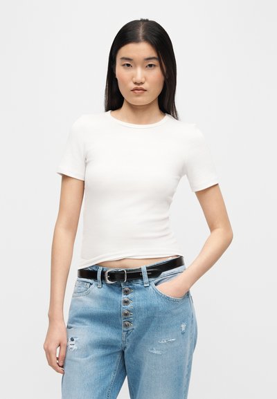 Νεαρή γυναίκα με μακριά σκούρα μαλλιά που φοράει λευκό κοντομάνικο πουκάμισο και ανοιχτό μπλε τζιν με σκισίματα και μαύρη ζώνη, με το χέρι στην τσέπη.