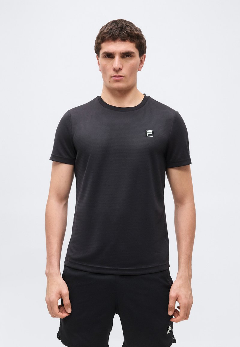 Άνδρας που φοράει μαύρο κοντομάνικο μπλουζάκι και μαύρο σορτς, στέκεται μπροστά σε απλό λευκό φόντο, κοιτάζοντας ευθεία με ουδέτερη έκφραση.