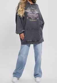 Femme portant un sweat à capuche noir délavé surdimensionné avec poche frontale, jeans à jambes larges bleu clair et baskets blanches, debout sur un fond uni.