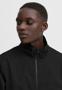 Chaqueta negra con un cuello alto y un cierre frontal. Textura de tejido suave, diseño minimalista, sin patrones ni acentos visibles.