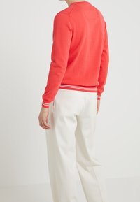 Roter Baumwollpullover mit weißen gestreiften Rippbündchen und Saum, kombiniert mit weißen Hosen. Die Rückseite zeigt ein einfaches Design mit einer Nahtdetails.