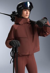 Rjav smučarski jopič iz gladkega materiala, v kombinaciji z oprijetimi rjavimi hlačami. Črni rokavici držita smučarske palice, črna čelada s temnimi očali pa zaključuje videz.