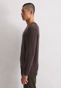 Mann in braunem Langarmpullover und dunkler Hose, der seitlich an einer schlichten weißen Wand steht.