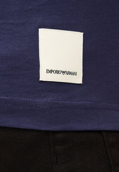 Κοντινό πλάνο ενός λευκού διακριτικού με το λογότυπο Emporio Armani ραμμένου στο τελείωμα ενός μπλε ενδύματος πάνω από μαύρο ύφασμα.