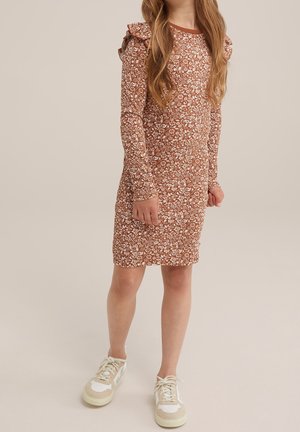 Ung pige iført en brun kjole med lange ærmer, knælængde og hvide blomsterprint samt hvide sneakers, stående mod en beige baggrund.