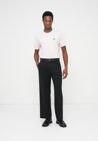 Hellrosa Kurzarm-T-Shirt mit gelbem Logo, kombiniert mit schwarzen maßgeschneiderten Hosen und schwarzen Lederschuhen. Einfaches Design und Passform.