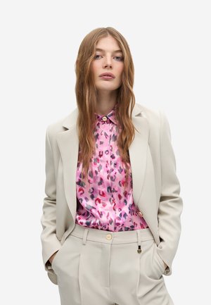 Chaqueta de traje beige ajustada sobre una blusa de leopardo rosa. La blusa tiene un collar y botón, con una textura de tela suave y brillante.