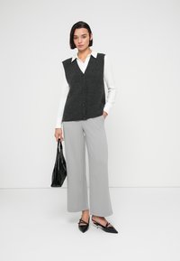 Σκούρο γκρι πλεκτό γιλέκο με V-λαιμόκοψη και κούμπωμα με κουμπιά, συνδυασμένο με ανοιχτό γκρι παντελόνι φαρδιάς γραμμής και μαύρα παπούτσια με μυτερή μύτη, κρατώντας μια μαύρη τσάντα χειρός.