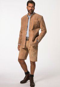 Giacca scamosciata beige con colletto alto e bottoni decorativi, abbinata a pantaloni corti fino al ginocchio con dettagli floreali ricamati. Stivali marroni.