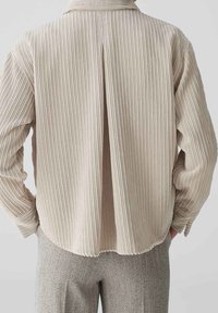 Chemise en velours côtelé beige clair avec un col, des rayures verticales et une fente au dos. Le tissu présente une texture et un ourlet arrondi.