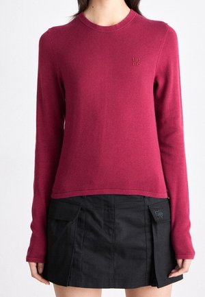 Žena nosící přiléhavý vínově červený svetr s dlouhým rukávem, doplněný černou mini sukní s předními klopovými kapsami.